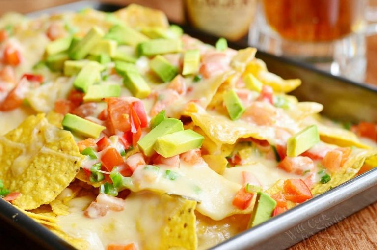 Close up view of Nachos with veggies and homemade nacho ale cheese on a silver baking pan on a wooden table with beer in the background