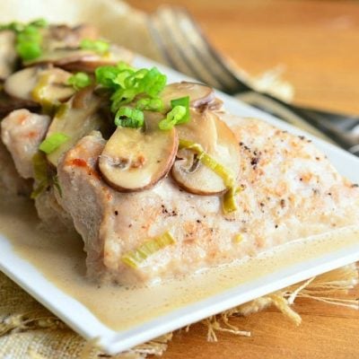 close up view of a white rectangular plate with creamy pork marsala entree sitting on a burlap cloth on a wooden table with a fork in the background to the right of the plate.