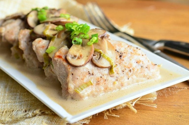 close up view of a white rectangular plate with creamy pork marsala entree sitting on a burlap cloth on a wooden table with a fork in the background to the right of the plate.