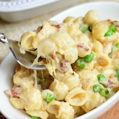 white rectangular baking dish filled with creamy white macaroni cheese with peas onions and bacon on a tan placemat with a tan cloth in the background and a small dish filled with the Macaroni cheese on a wooden table and a spoon taking a portion out