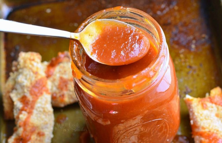 mason jar filled with homemade whiskey BBQ sauce standing on a baking pan with easy baked chicken strips and a spoon lifting some sauce above the jar