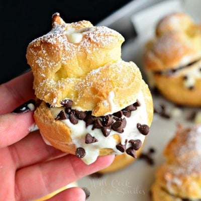 hand holding on cannoli above a Baking pan with wax paper holding cannoli pastry cream puffs as viewed from above on a black table