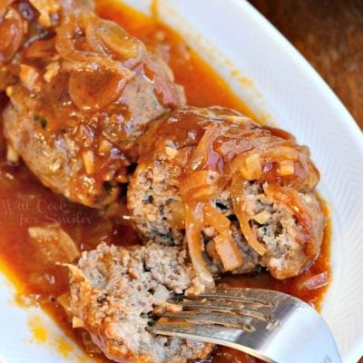 small white oval dish with french onion baked meatballs on a wooden table with a fork splitting the first meatball and taking one bite