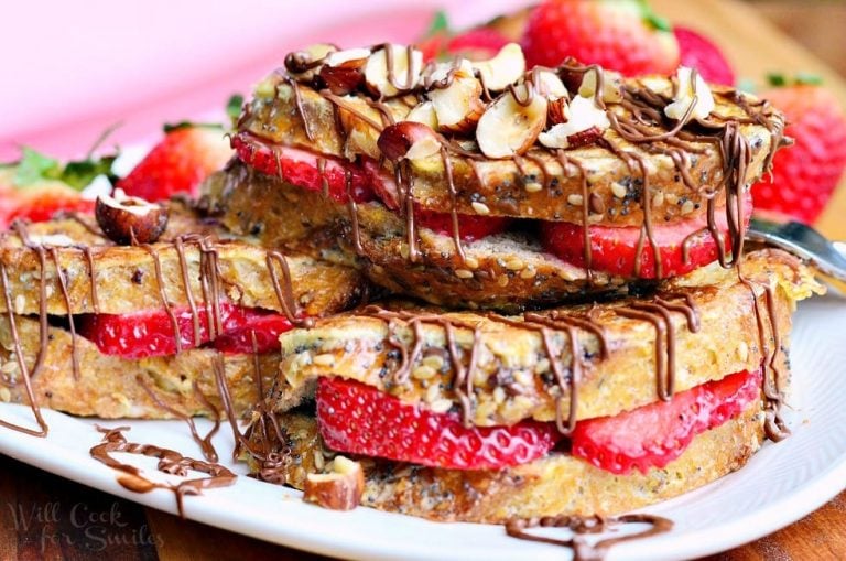 Small round white plate with Fruit filled hazelnut french toast with chocolate drizzle topping on a wooden table with a fork in the foreground and a pink cloth in the background to the left