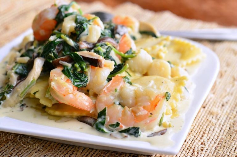 White decorative plate with ravioli with seafood spinach mushrooms in garlic sauce on a tan placemat on a wooden table with a spoon in the background.