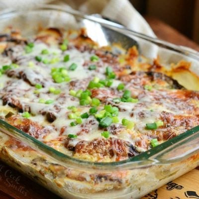 Glass baking dish with scallopred potatoes portobello au gratin on a wooden cutting board with a silver spoon and whiteish tan cloth in the background