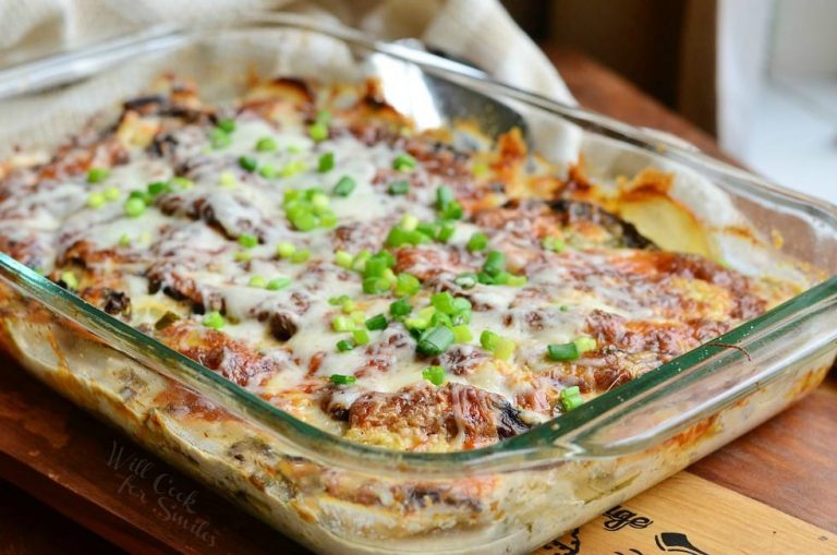 Glass baking dish with scallopred potatoes portobello au gratin on a wooden cutting board with a silver spoon and whiteish tan cloth in the background