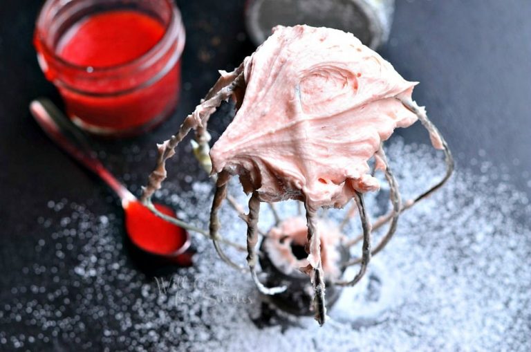 mixer attachment standing on a black table covered in powdered sugar while the attachment is covered in strawberry buttercream frosting as viewed from above