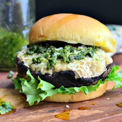 front view of Italian stuffed portabello burger on a bun and bed of lettuce sitting on a wooden board with a spoon of pesto at the bottom left and block of cheese in the background along with a bottle of pesto