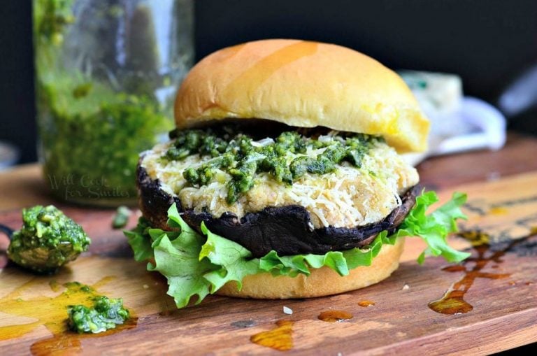 front view of Italian stuffed portabello burger on a bun and bed of lettuce sitting on a wooden board with a spoon of pesto at the bottom left and block of cheese in the background along with a bottle of pesto