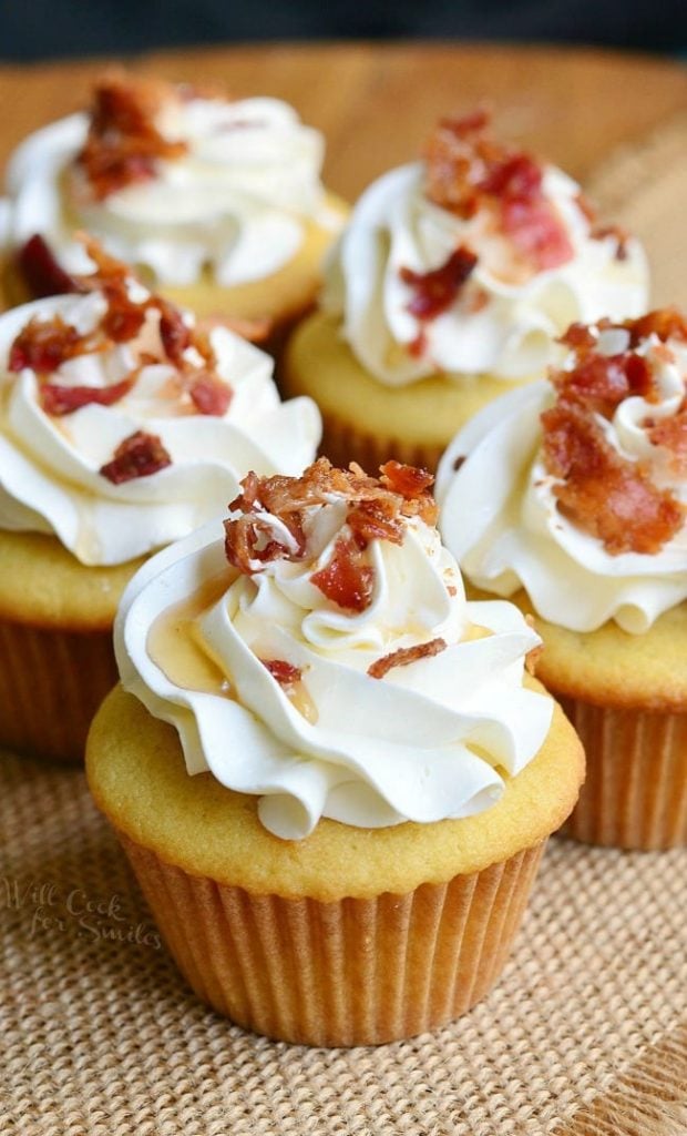 view from the front of 4 maple bacon cupcakes on a burlap cloth on wooden table with a black background