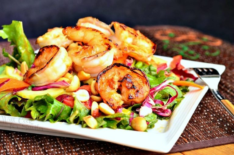 close up view from the front of thai shrimp salad with peanut dressing on a red bamboo placemat and salad is on a white square plate and a fork is leaning on the right side of the plate