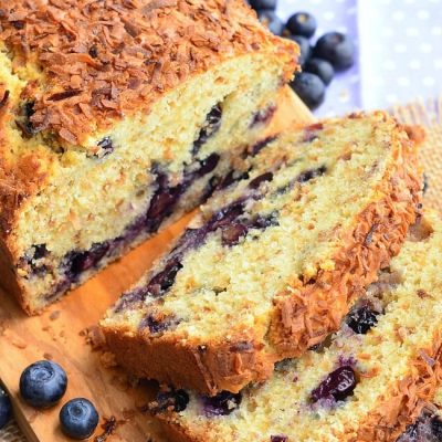 loaf of toasted coconut blueberry bread with the first 3 slices stacked in front of the rest of the loaf on a wooden cutting board with blueberries scattered around on table.