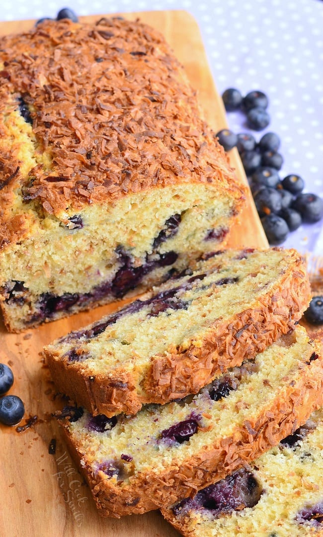 view from above of loaf of toasted coconut blueberry bread with the first 3 slices stacked in front of the rest of the loaf on a wooden cutting board with blueberries scattered around on table.