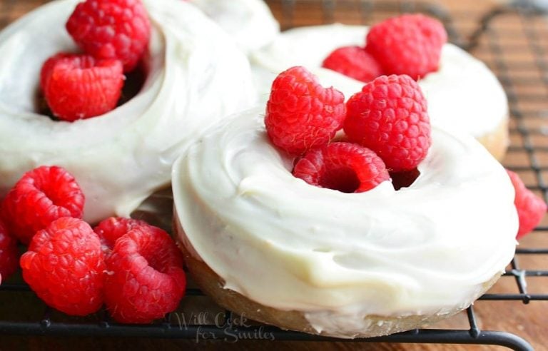 close up view of 4 white chocolate covered baked raspberry donuts on a wire cooling rack and whole raspberries laying at bottom left of donuts.