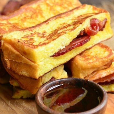 Cut bacon stuffed brioche french toast sticks stacked on a wooden cutting board with a small bowl of syrup