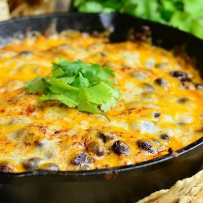 close up view of a black skillet with hot 7 layer dip rests on a table surrounded by chips redy for dipping.