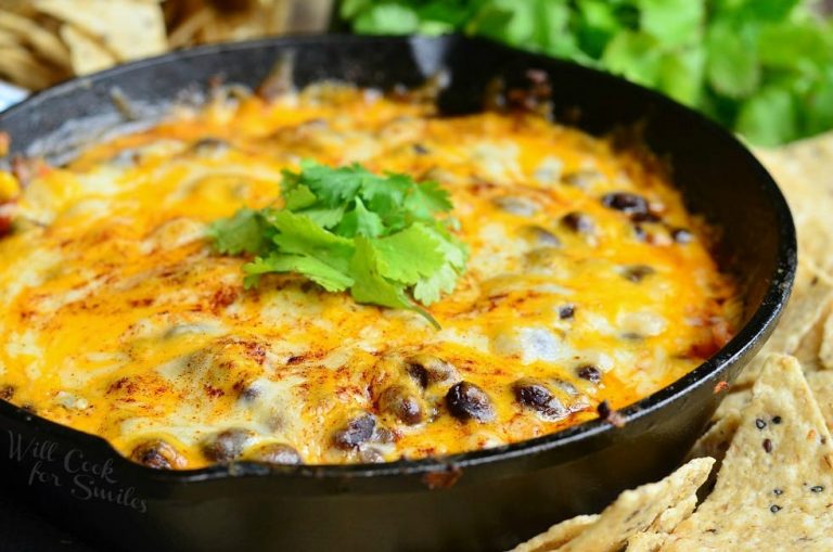 close up view of a black skillet with hot 7 layer dip rests on a table surrounded by chips redy for dipping.