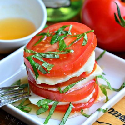 small white square plate with fresh caprese salad with a bottle of oil and additional tomatoes in the background