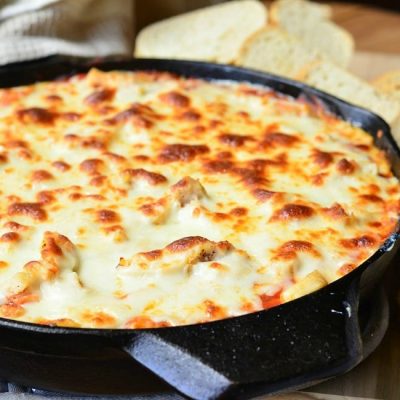 close up view of black skillet filled with chicken parmesan dip on a wooden table with sliced bread and a tan cloth in the background