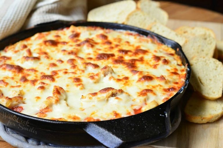 close up view of black skillet filled with chicken parmesan dip on a wooden table with sliced bread and a tan cloth in the background