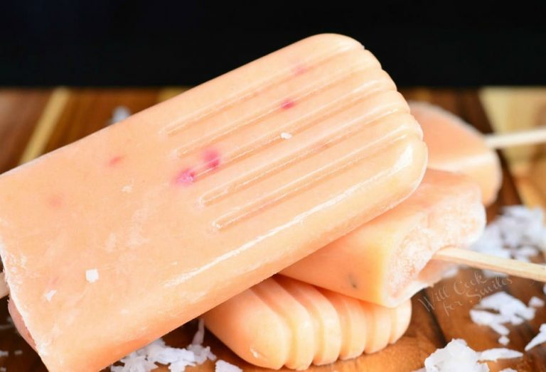 close up view of 3 coconut and peach creamy popsicles on a wooden table with coconut shaving scattered in front of popsicles