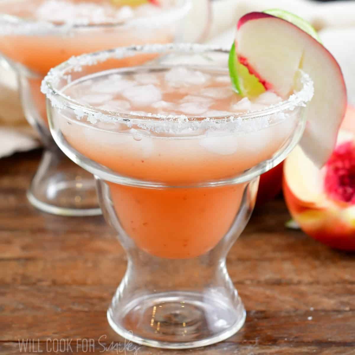 peach margarita in a glass with peaches around and another glass behind.