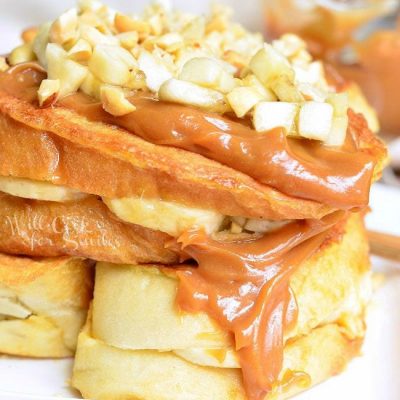 white square plate with peanut butter banana stuffed french toast and a fork laying in the corner of the plate all on a wooden table with a glass sauce boat in the background