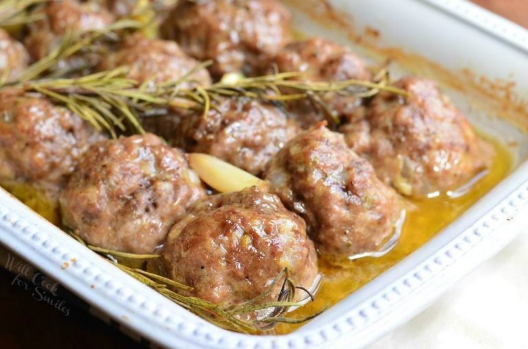 close up view of roasted garlic rosemary baked meatballs in a white baking dish on a wooden table topped with 3 rosemary sprigs