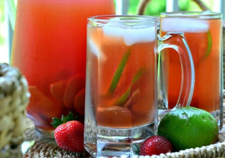 Clear glass beer mug filled with strawberry lime infused iced tea in front of another mug and pitcher filled with tea viewed close up