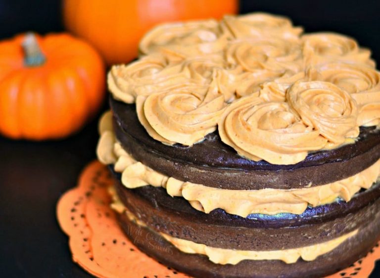 close up view of chocolate cake with pumpkin cream cheese frosting on orange doilies with 2 pumpkins in the background