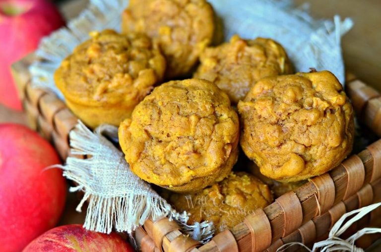 wooden basket filled with pumpkin apple muffins on a wooden table with red apples placed around the basket viewed close up