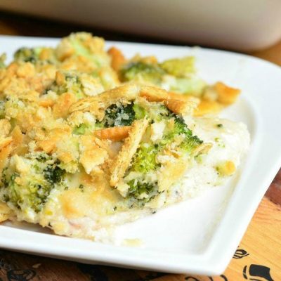 white decorative square plate with 1 portion of broccoi cheese chicken and a white baking dish filled with the rest of the entree in the background as viewed close up