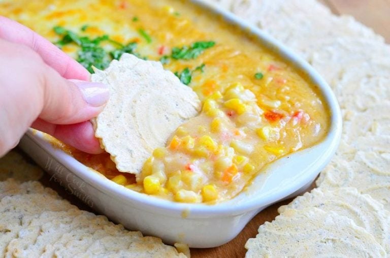 corn chowder hot dip in a white oval dish with crackers around the dish while all sit on a wooden table and a hand dips one cracker into dip viewed close up