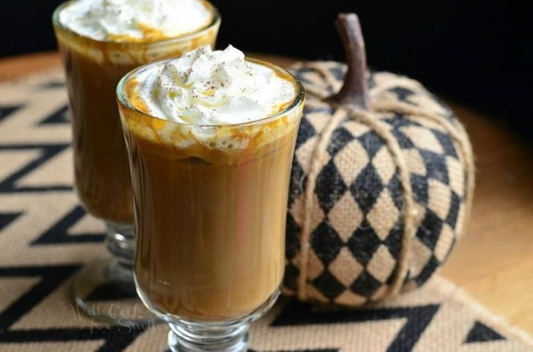 Dessert coffee glass mug filled with pumpkin white chocolate mocha latte on a white and black zig zag placemat with an additional glass in the background to the left next to a fabric pumpkin decoration