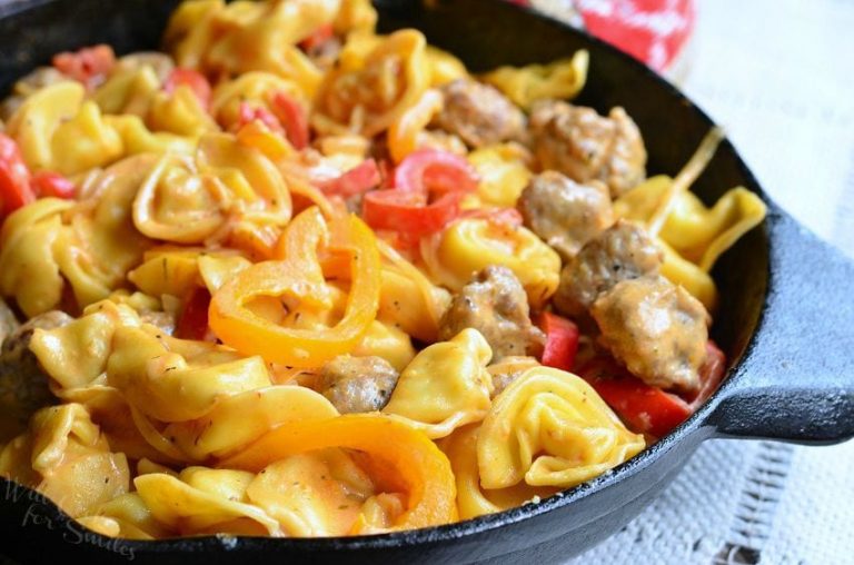 black skillet filled with sausage peppers and onion tortellini dish on a light blue cloth with a red and white cloth in the background as viewed close up