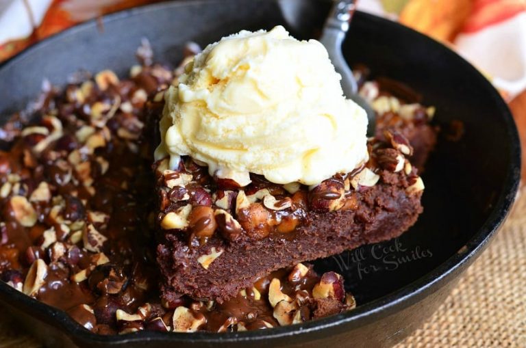 1 slice of brownie being held above black skillet filled with a small batch of pumpkin turtle brownie on a wooden table as viewed close up