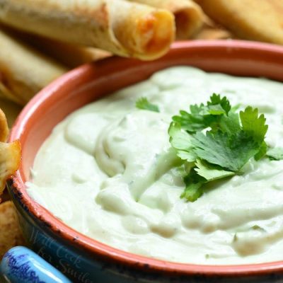 Avocado ranch dip in a blue and orange crock with taquitos leaning on crock