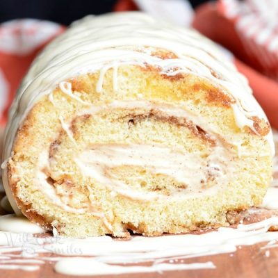 View from the front of cinnamon roll cake roll on a wooden board with icing drizzled across the top of the roll and a white and red cloth in the background