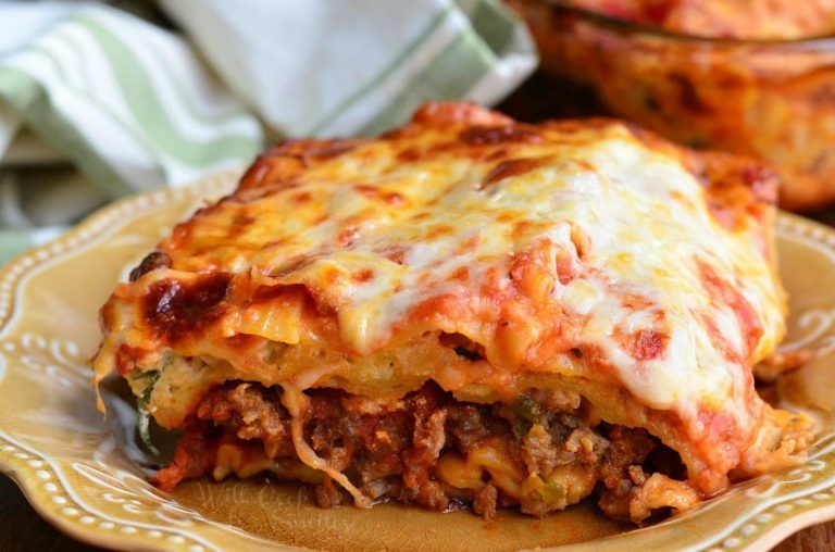 1 slice of Italian sausage lasagna on a light yellow colored plate on a wooden table with the rest of the lasagna in a clear baking dish in the background to the right