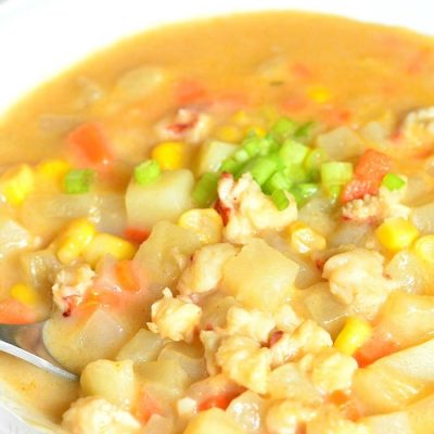 view from above of a white bowl filled with lobster potato and corn chowder as a spoon rests on the bowl and a brown cloth sits in the background