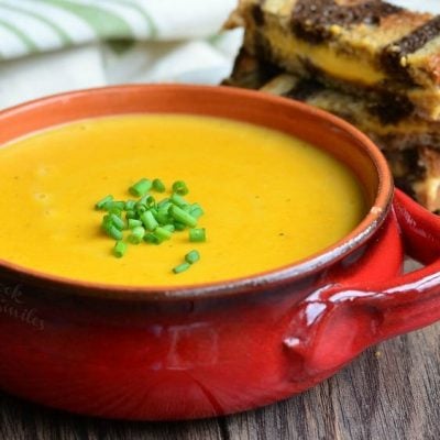 Red clay crock filled with pumpkin ale soup on a wooden table with a cut up grilled cheese in the background and a white and green cloth in the background to the left