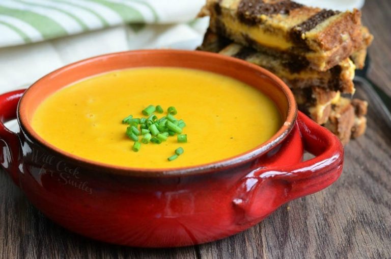 Red clay crock filled with pumpkin ale soup on a wooden table with a cut up grilled cheese in the background and a white and green cloth in the background to the left