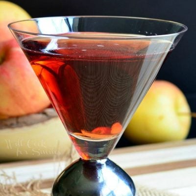 Side view of a decorative martini glass with washington apple martini on a brown placemat with a bowl of apples in the background to the left