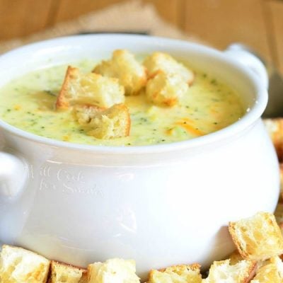 White crock filled with asiago cheese soup on a yellow plate with cut bread croutons scattered around the bowl placed on the plate on a black and white placemat on a wooden table