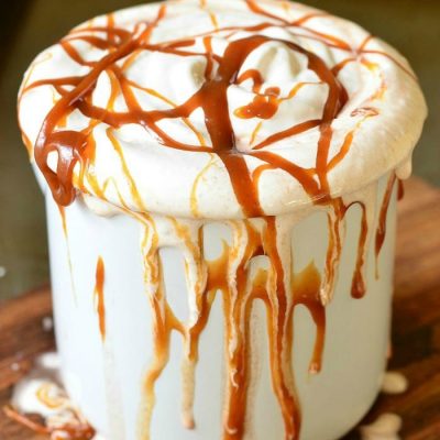 Metal dessert mug filled with caramel marshmallow hot chocolate on a wooden table covered in caramel drizzle