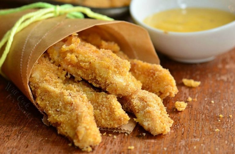 Wax paper holder holding easy honey mustard baked chicken fries on a wooden table with a small white saucer in the background filled with sauce and a pan with additional chicken fries in the background to the left as viewed close up