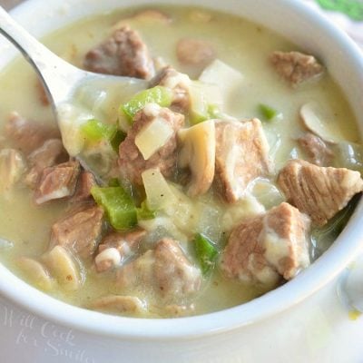 white crock filled with steak and cheese soup on a wooden table with a spoon holding up one bite of soup above the crock