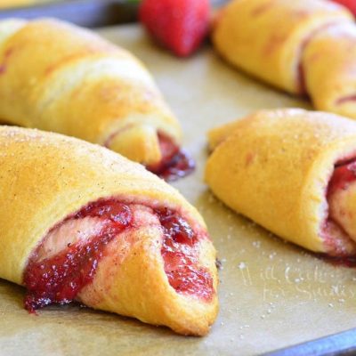 6 strawberry crecent rolls on a baking sheet tray with strawberries scattered around the tray as viewed close up