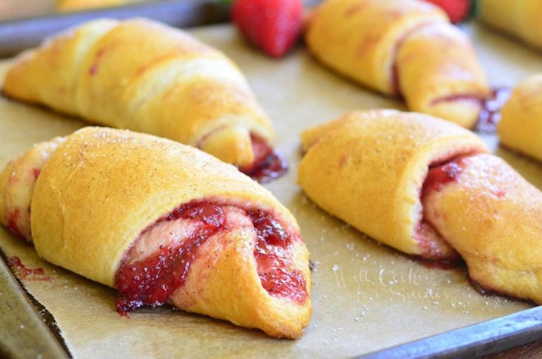 6 strawberry crecent rolls on a baking sheet tray with strawberries scattered around the tray as viewed close up